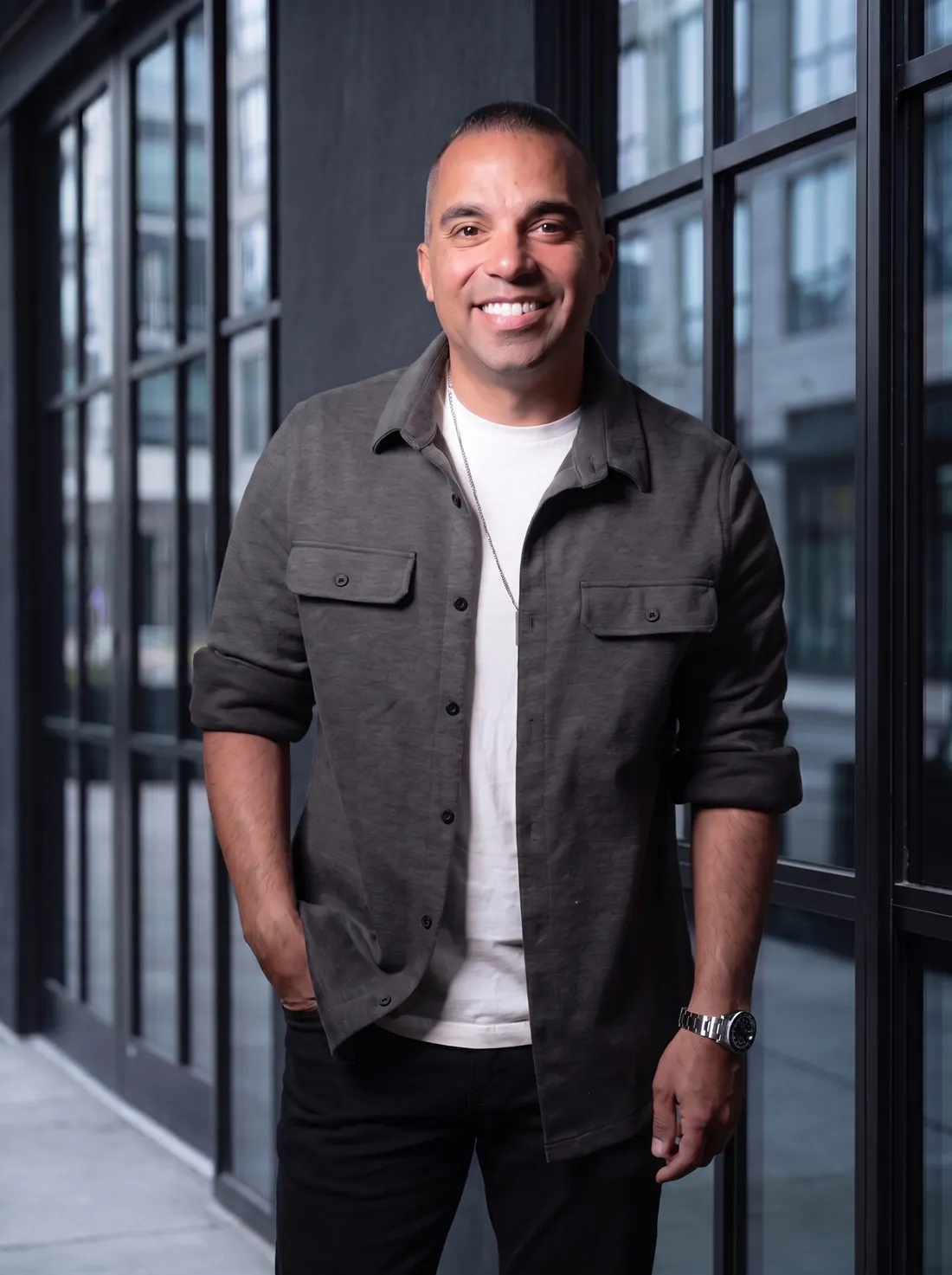 Tyler Perez smiling in front of black steel-framed windows in a charcoal shirt jacket